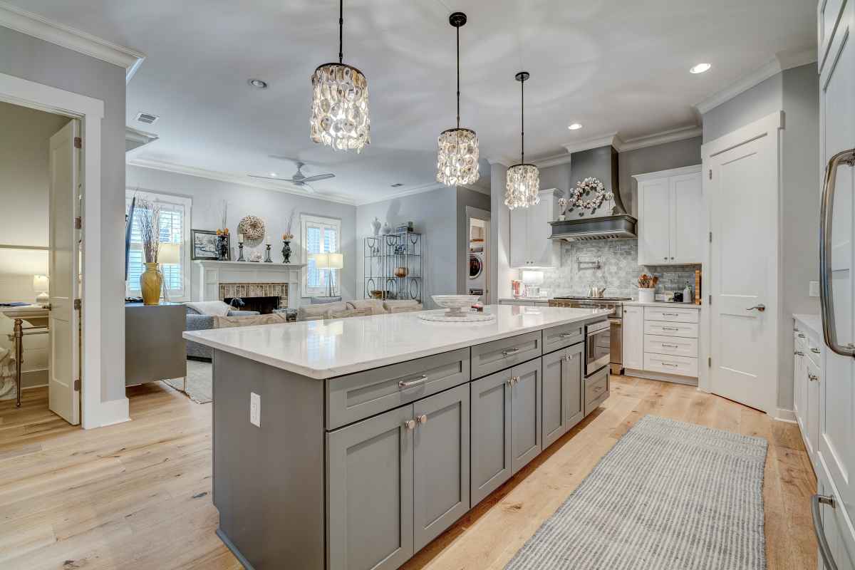 Modern kitchen with gray cabinets, large island with white countertop, three pendant lights, hardwood flooring, and adjoining cozy living room.