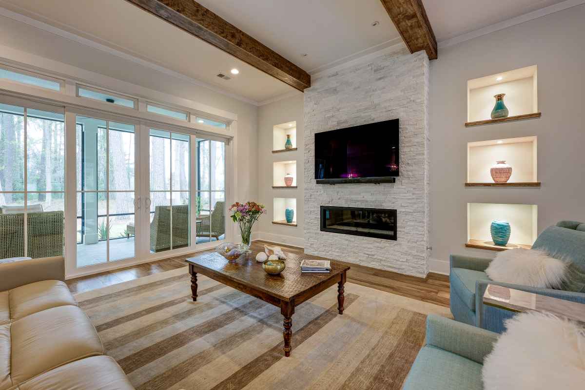 A cozy living room with a striped rug, beige sofa, and blue armchairs. A wall-mounted TV and fireplace are framed by shelves with colorful vases, and large windows overlook a patio.