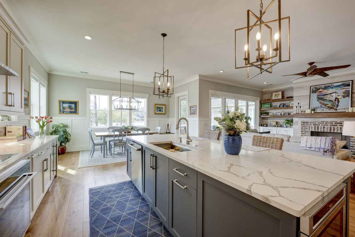 Spacious kitchen with a large marble island, gray cabinets, and pendant lighting. Dining area and cozy living room with shelves in the background.