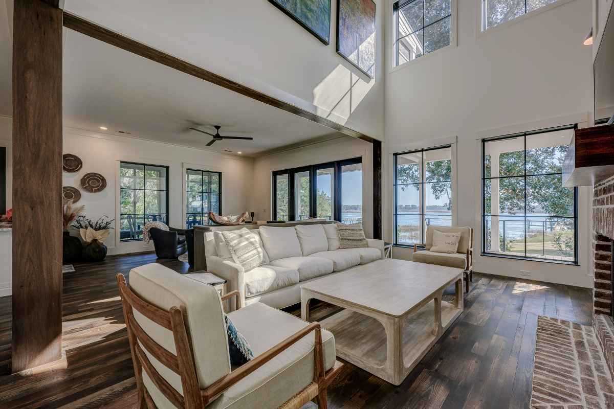Sunny living room with high ceilings features a beige sofa set, wooden floors, large windows with lake views, and a modern ceiling fan. Airy and serene atmosphere.