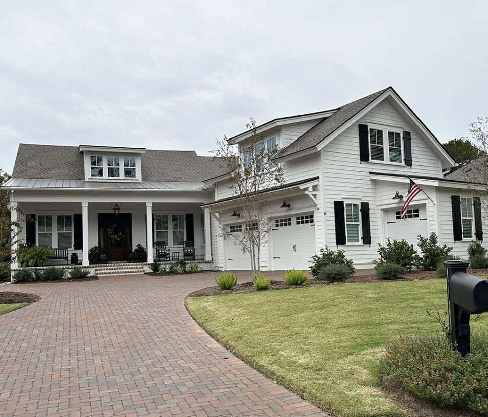 A white, two-story suburban house with dark shutters and a gray roof. It features a front porch, an American flag, and a brick driveway, surrounded by greenery.