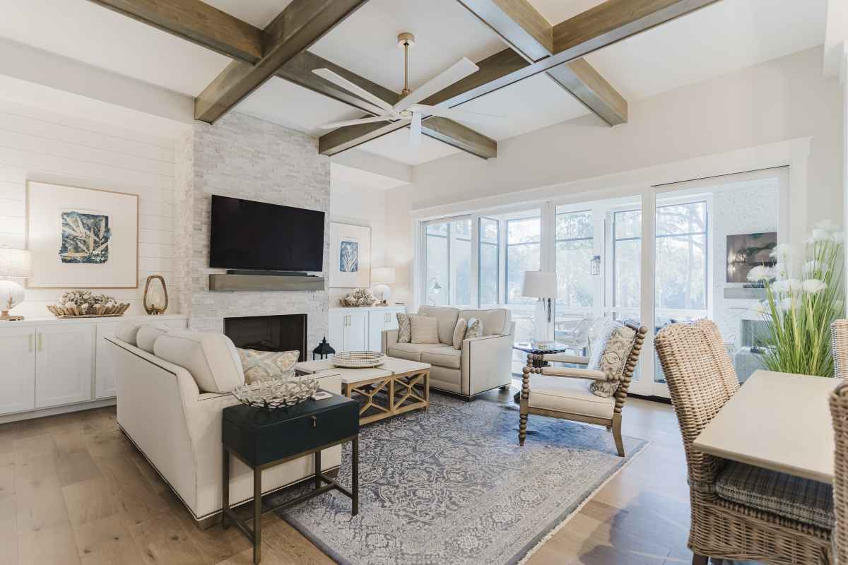 Chic living room with wooden beams, gray sofas, a blue rug, and a stone fireplace. Large windows provide natural light.