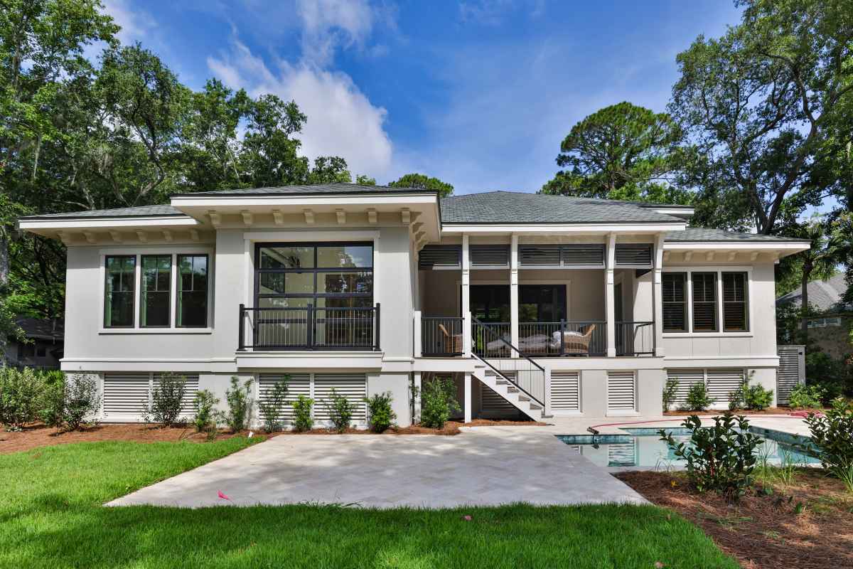 The image shows the rear exterior of a modern, single-story home with a patio, swimming pool, and screened-in porch. Lush green trees surround the property under a bright blue sky.