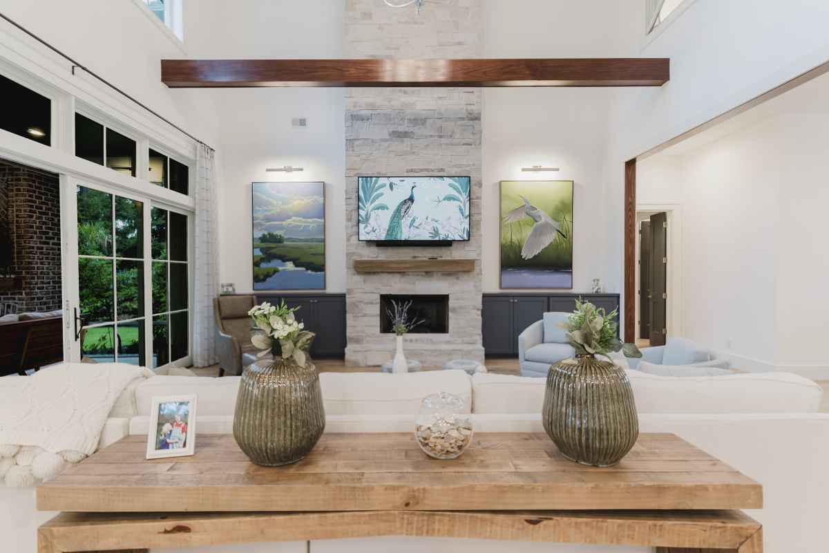 Modern living room with a high ceiling and neutral tones. Features a stone fireplace with a TV above, large artwork, wide windows, and cozy seating.