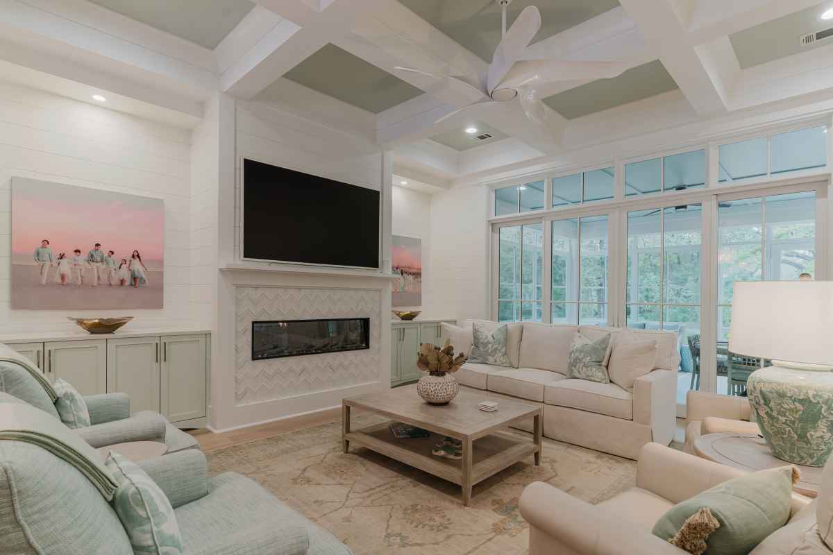 A bright and airy living room features comfortable seating, a modern fireplace and large windows leading to an outdoor space. A family portrait hangs above a credenza on the left side of the room.