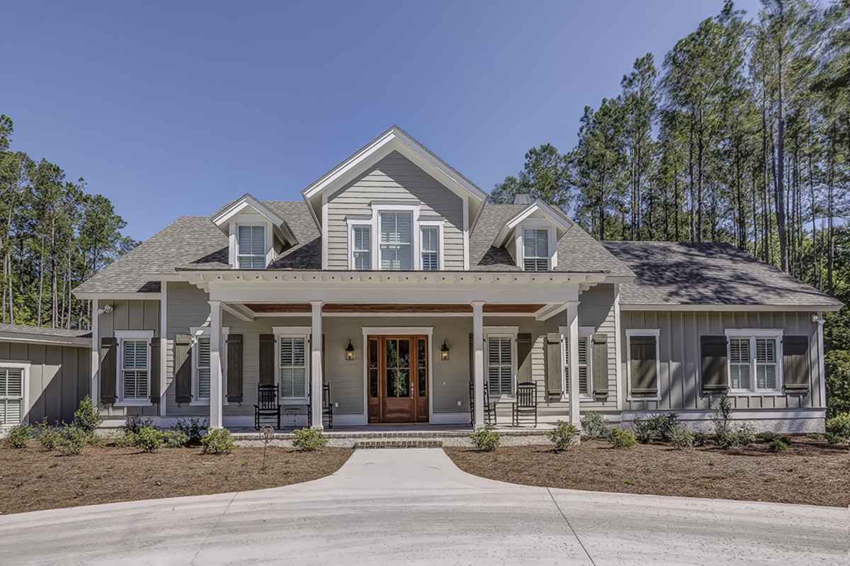 A charming modern farmhouse with light gray siding, large windows, and a covered porch with rocking chairs, set against a background of tall pine trees.