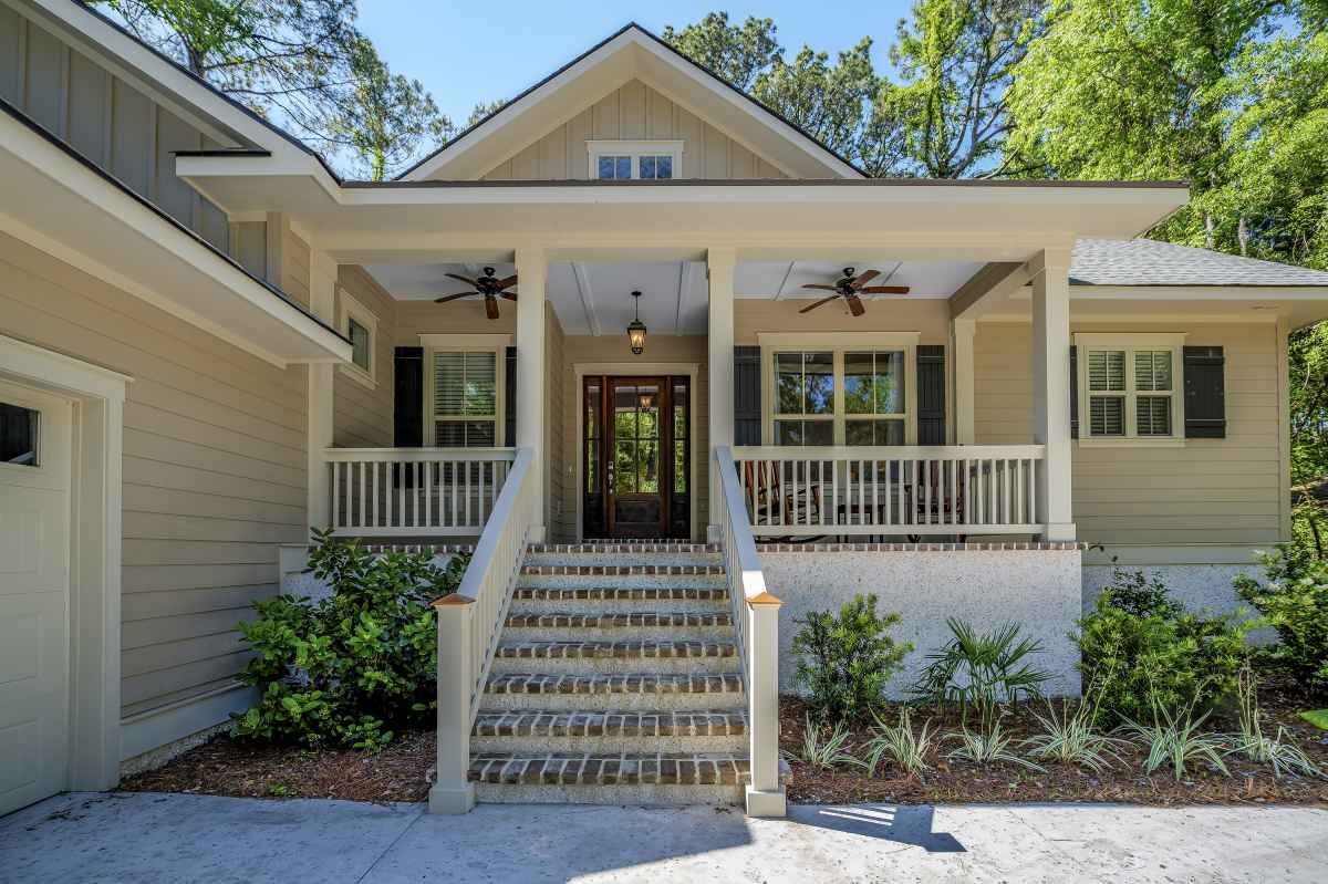 Charming, beige house with a front porch, steps leading up, ceiling fans, white railings, and surrounding greenery under a clear blue sky.