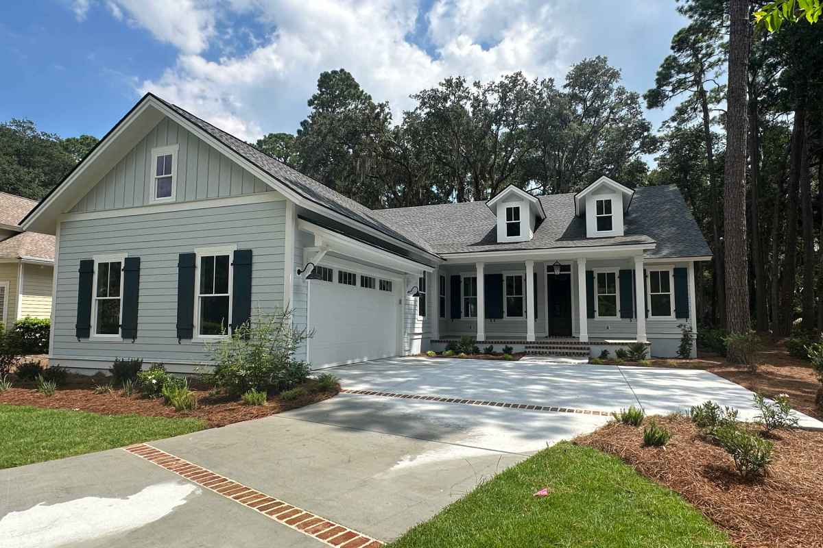 Charming single-story house with light blue siding, dark shutters, and a covered porch. Surrounded by trees and a well-maintained lawn under a blue sky.