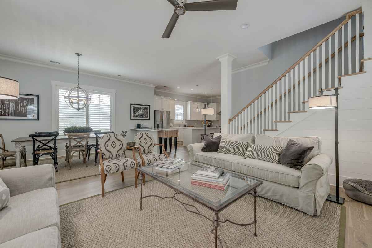 Bright, modern living room with neutral tones featuring two sofas, a glass coffee table, patterned chairs, and a dining area under a stylish pendant light.
