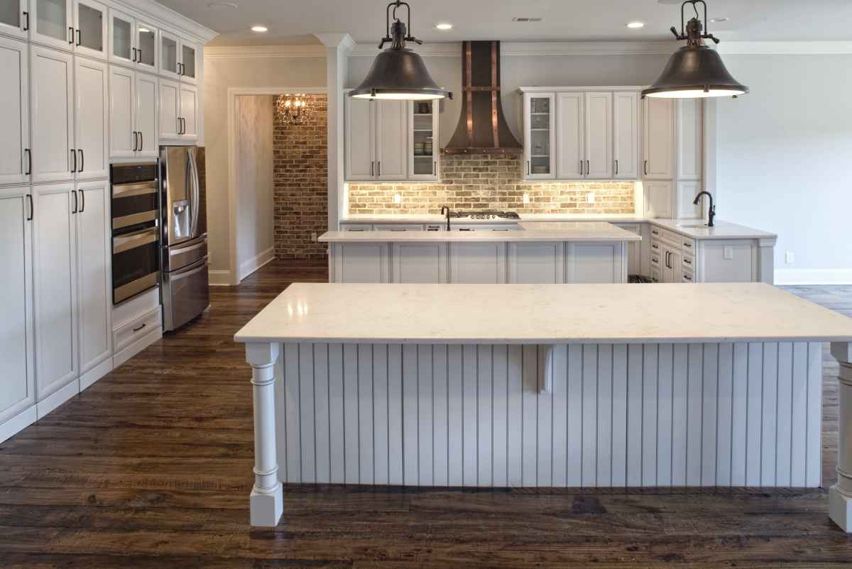 Spacious kitchen with rustic wooden floors, white cabinetry, marble countertops, two large pendant lights, and exposed brick backsplash, conveying elegance.