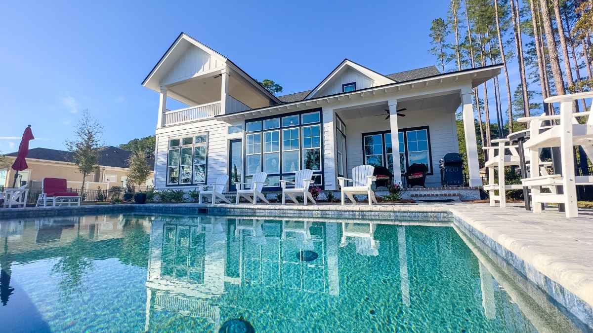 Modern house with large windows overlooking a clear, aqua blue pool. Adirondack chairs and trees in the background convey a serene, vacation-like atmosphere.