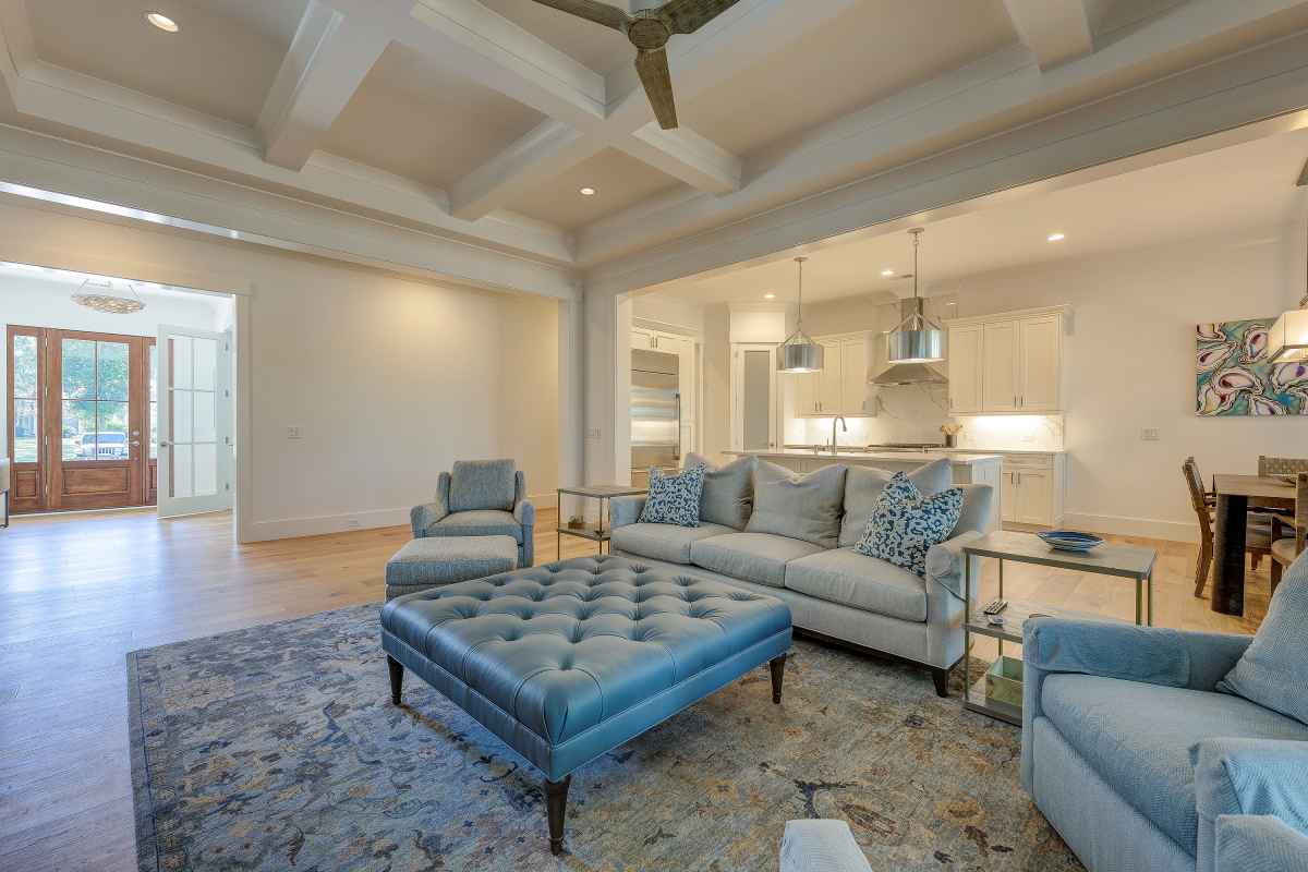 Elegant living room with plush blue seating, tufted ottoman, and cozy rug. Open layout leads to a bright kitchen and dining area.