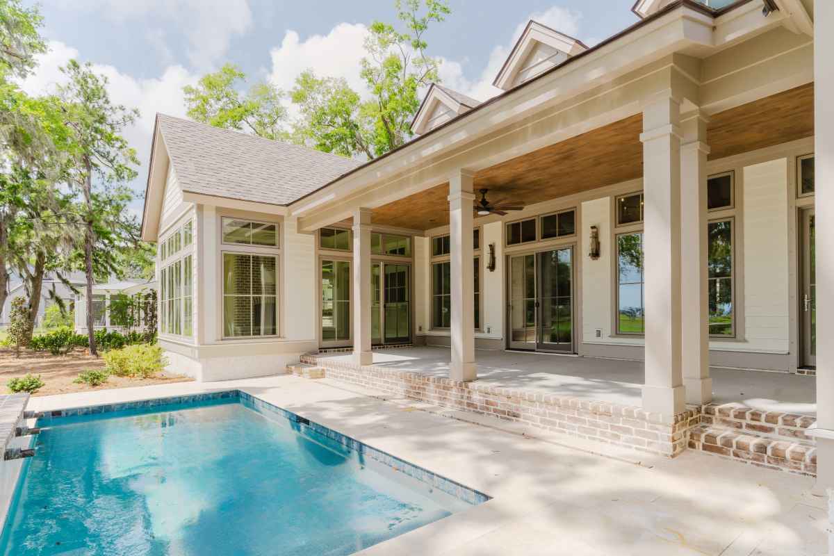  spacious modern house with large windows and a covered patio overlooks a small, leaf-strewn pool. Surrounded by lush green trees on a sunny day.