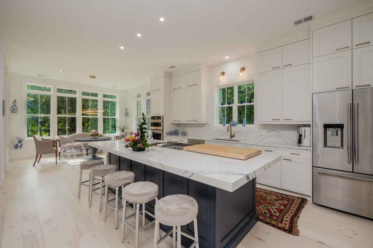 A bright and modern kitchen features a large island with seating, white cabinets and stainless steel appliances. The space opens up to a dining area with large windows overlooking greenery.