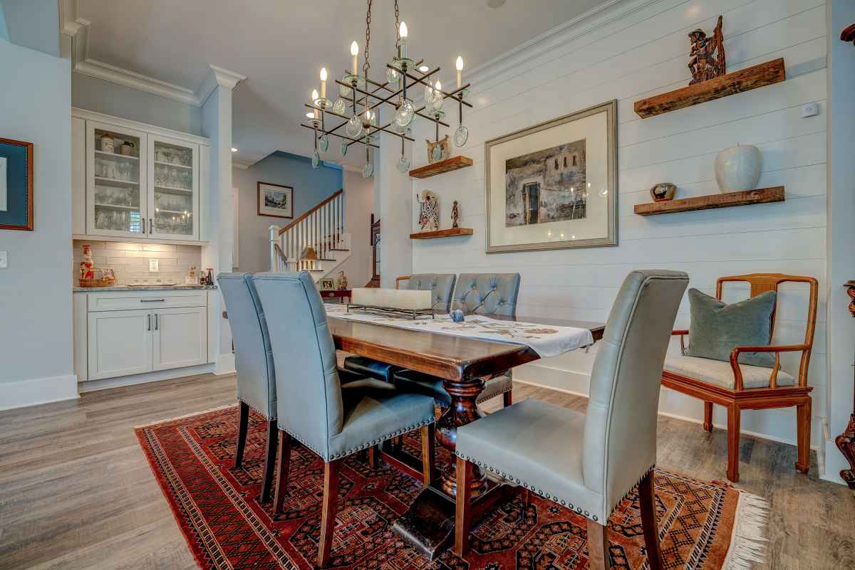 A cozy dining room features a wooden table with gray cushioned chairs on a red patterned rug. A chandelier hangs above, with art and shelves on the walls.
