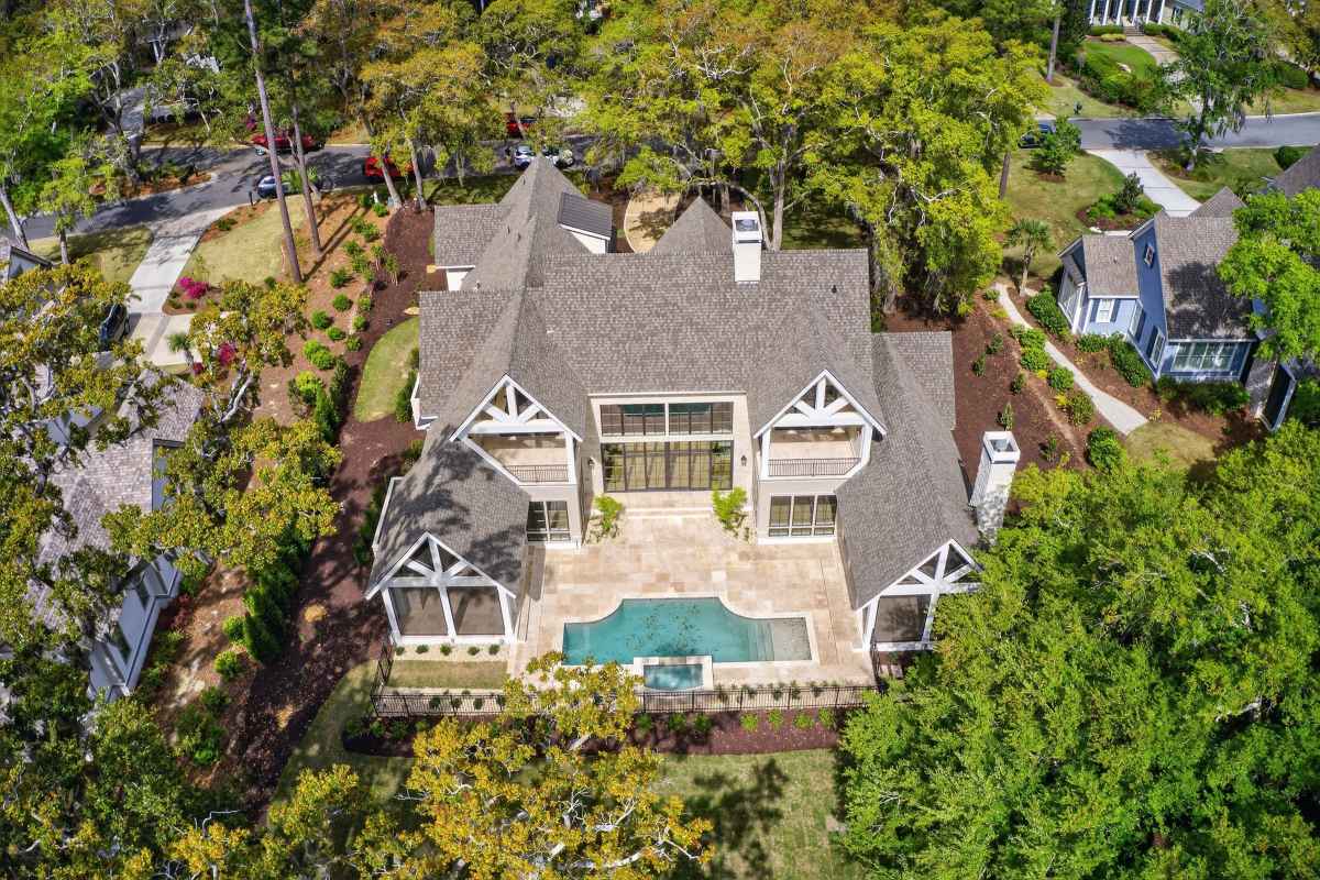 A rustic, two-story stone house with large windows, surrounded by lush greenery and colorful flowers, under a clear blue sky.
