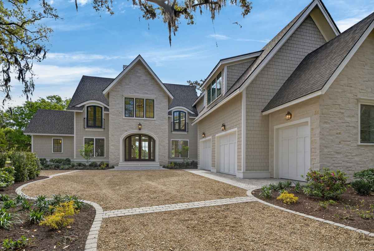 A rustic, two-story stone house with large windows, surrounded by lush greenery and colorful flowers, under a clear blue sky.