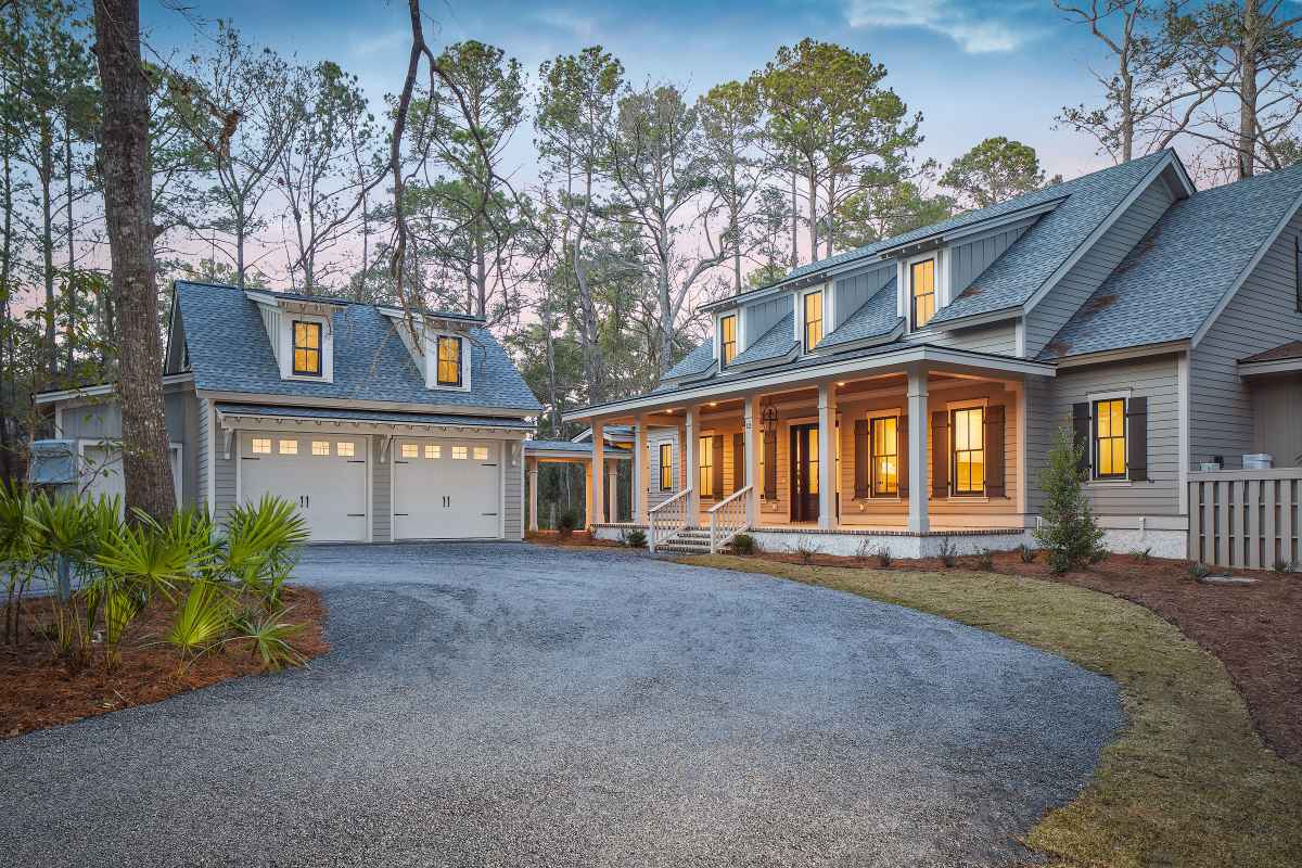 A cozy two-story house with lit windows and a large front porch sits under twilight sky, surrounded by tall trees, with an inviting driveway and garage.