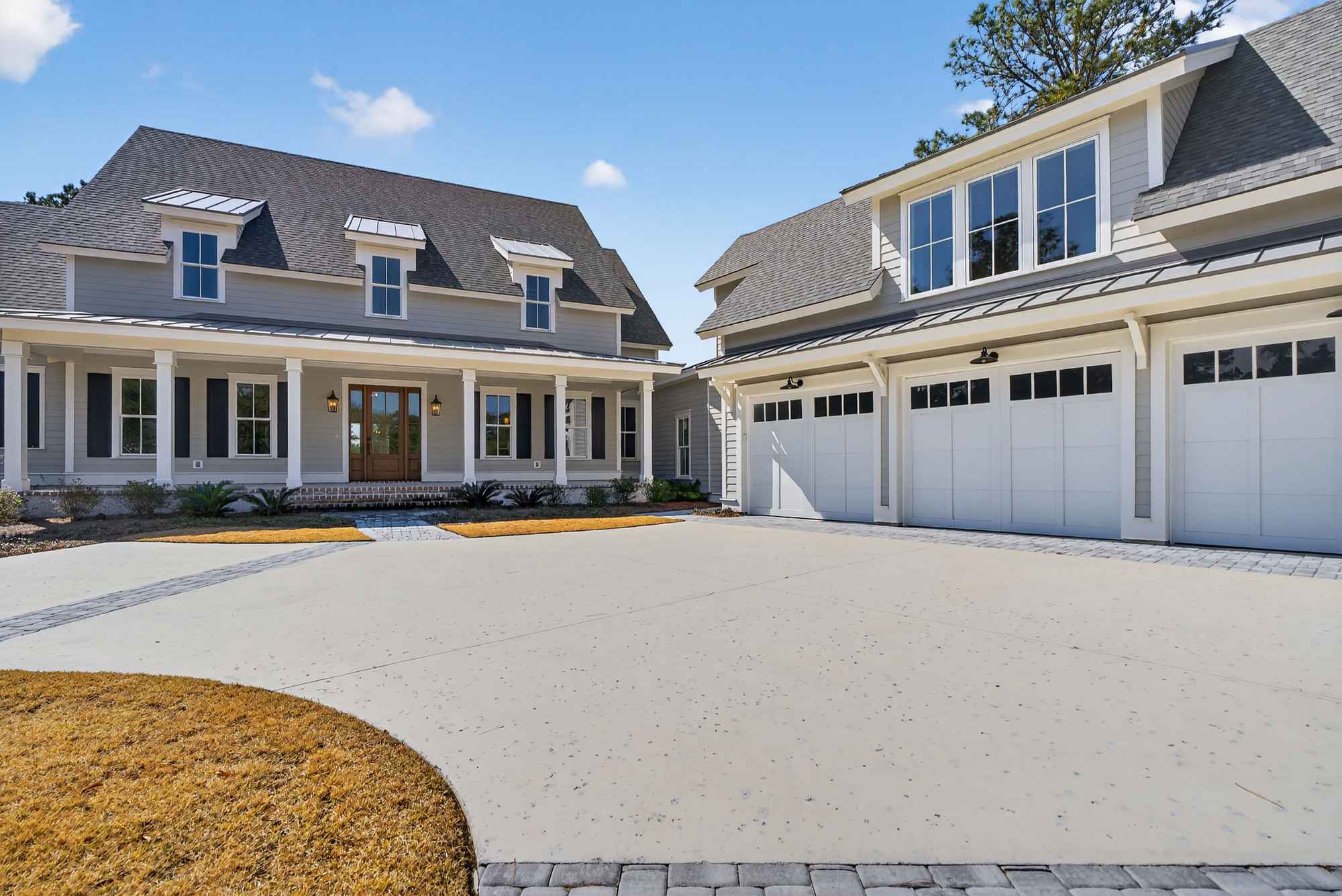 A charming two-story house with a wraparound porch, adjacent garage, and surrounded by lush greenery, set against a backdrop of tall pine trees, under a clear blue sky.